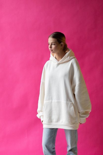 Beautiful blonde woman in a white hoodie and blue jeans posing on a pink background