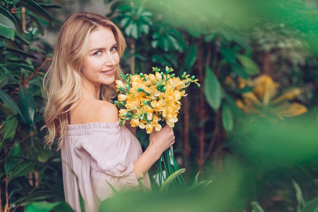 Free photo beautiful blonde young woman standing near the plants holding yellow freesia