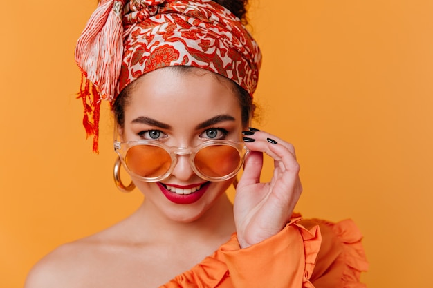 Free photo beautiful blue-eyed lady in unusual headband and red lipstick takes off sunglasses and poses on orange space.