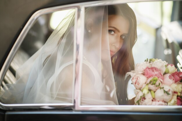 Beautiful bride sits with wedding bouquet in a retro car and has fun