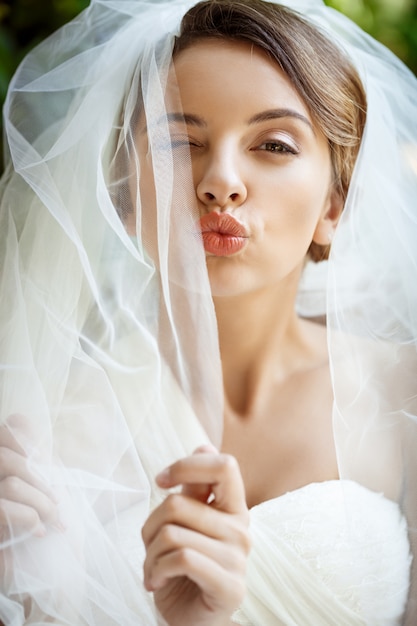 Free photo beautiful bride in wedding dress and veil winking, sending kiss.