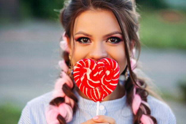 Beautiful brunette girl hiding face by candy heart on stick.