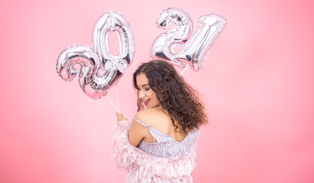 Beautiful brunette girl with bare shoulders in festive clothes posing on a pink background with balloons for the new year in her hands