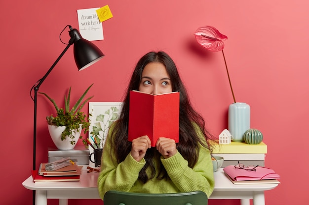 Free Photo beautiful brunette pensive mixed race female student learns information from textbook, covers half of face with red diary