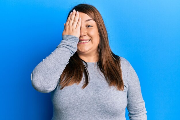 Beautiful brunette plus size woman wearing casual clothes covering one eye with hand, confident smile on face and surprise emotion.