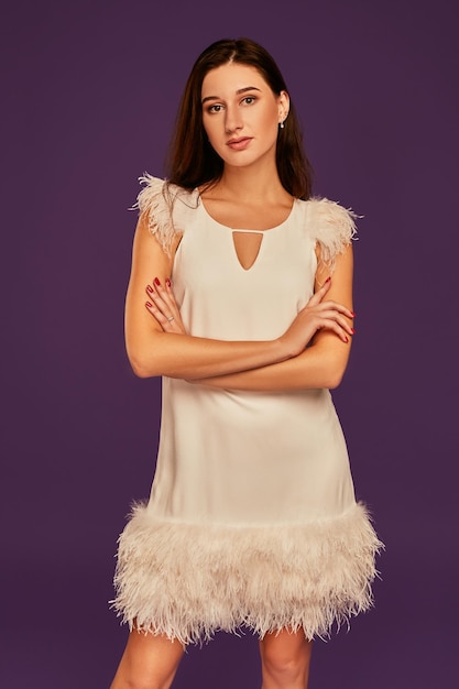 Free Photo beautiful brunette woman in elegant cocktail white dress is posing in studio on purple background. three quarter length studio shot