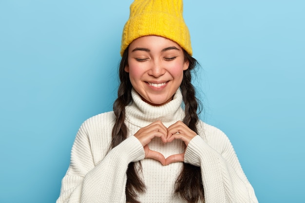 Free photo beautiful brunette woman keeps eyes closed with pleasure, makes heart gesture, expresses her true love, dressed in casual white sweater