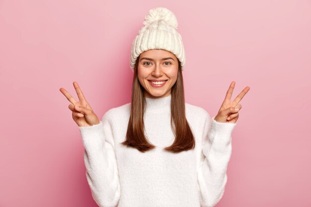 Beautiful brunette woman shows victory or peace gesture, smiles pleasantly, being in high spirit, wears white hat with pompon and sweater, isolated over pink wall