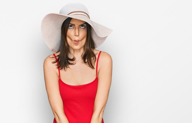 Beautiful brunette woman wearing swimsuit and summer hat making fish face with lips, crazy and comical gesture. funny expression.