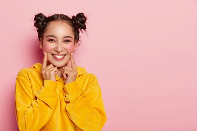 Beautiful brunette woman with two buns, points index fingers on cheeks, dressed in casual corduoy yellow sweatshirt, wears bright pinup makeup