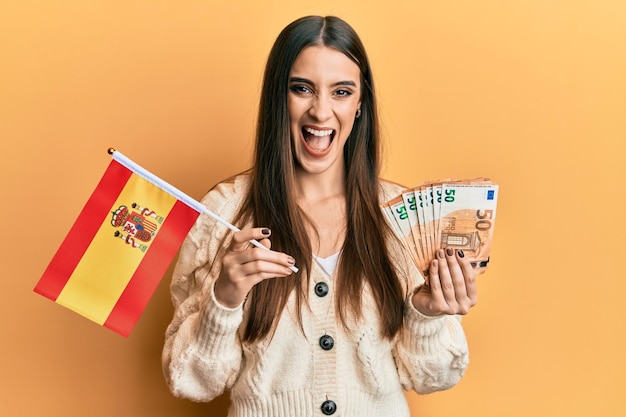 Free Photo beautiful brunette young woman holding spain flag and euros banknotes smiling and laughing hard out loud because funny crazy joke.