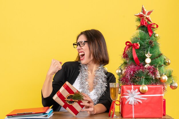 Beautiful business lady in suit with glasses holding her gift proudly sitting at a table with a xsmas tree on it in the office