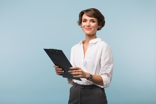 Free Photo beautiful business woman with dark short hair in white shirt holding black folder in hands while joyfully looking in camera over blue background