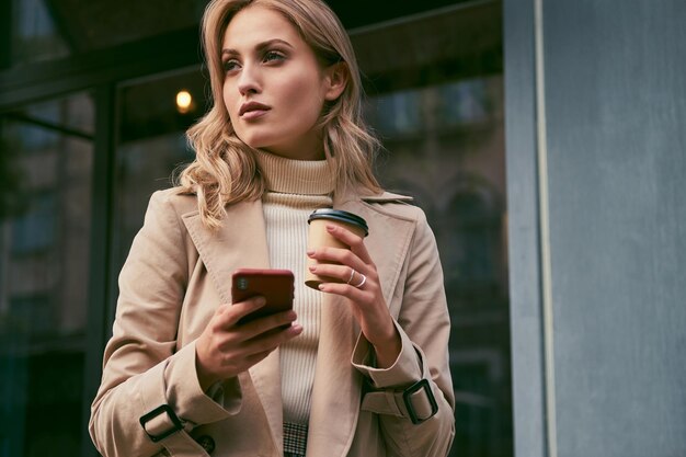 Beautiful casual blond girl in trench coat with coffee to go and cellphone thoughtfully looking away outdoor