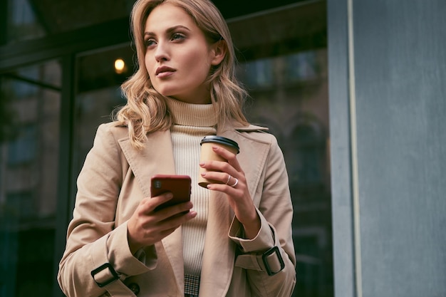 Beautiful casual blond girl in trench coat with coffee to go and cellphone thoughtfully looking away outdoor