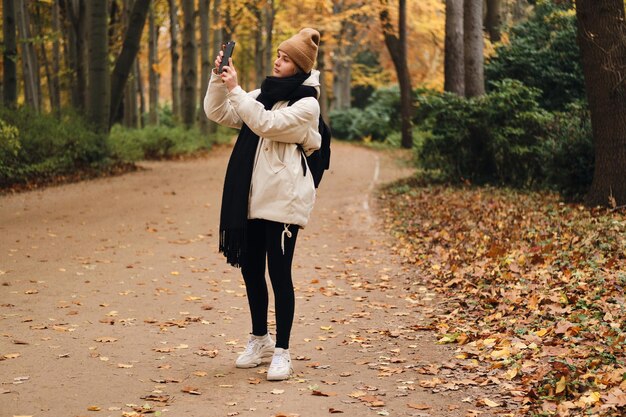 Beautiful casual girl walking through autumn park and taking photos on cellphone