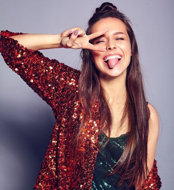 Beautiful caucasian smiling hipster brunette woman model in bright shinny reflecting summer stylish jacket and green dress posing on gray . Showing peace sign