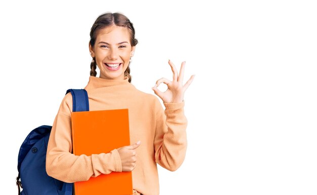 Beautiful caucasian woman with blonde hair wearing student backpack and holding book doing ok sign with fingers smiling friendly gesturing excellent symbol