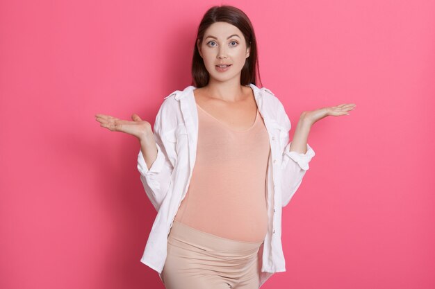 Beautiful cheerful european pregnant woman in comfy outfit spreading hands with surprised expression