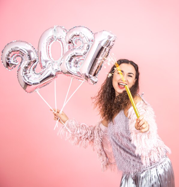 Beautiful cheerful festively dressed brunette girl with curly hair posing on a pink studio background with silver balloons for the new year concept