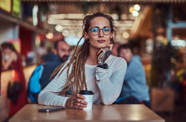 Beautiful cheerful girl with tattooes and dreadlocks is sitting at food court while drinking coffee.