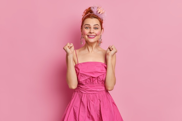 Free photo beautiful cheerful woman with red hair and make up clenches fists rejoices something dressed in festive dress and headband has happy expression poses