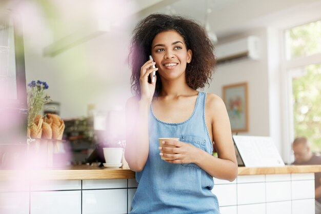 Beautiful cheerful young african woman student smiling talking on phone drinking coffee in cafe.