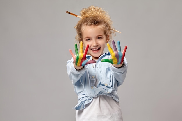 Beautiful child having a brush in her chic curly blond hair, wearing in a blue shirt and white t-shirt. She is showing her painted fingers and looking at the camera, on a gray background.