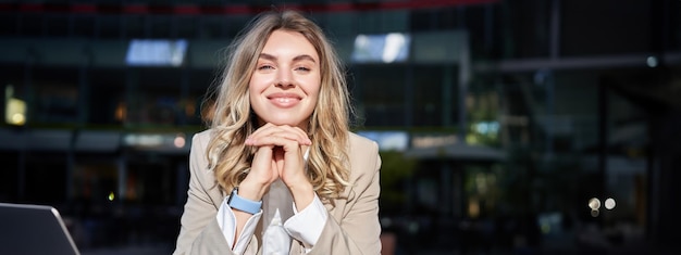 Free photo beautiful corporate woman looks dreamy and smiles stands outside on street leans her head on hands