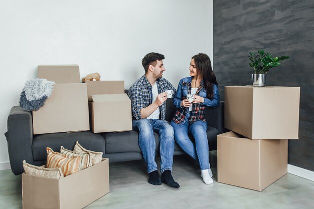 Beautiful couple in casual clothes is discussing plan of their new house and smiling while lying on the sofa near boxes for move