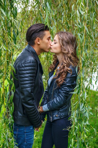 Beautiful couple in leather jackets kissing under green tree.