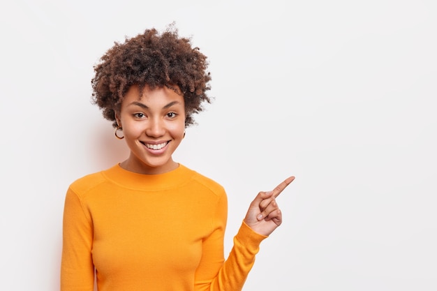 Beautiful curly haired Afro American woman suggests to clickon link points on blank copy space shows way stands over white wall wears basic orange jumper. People and advertising concept