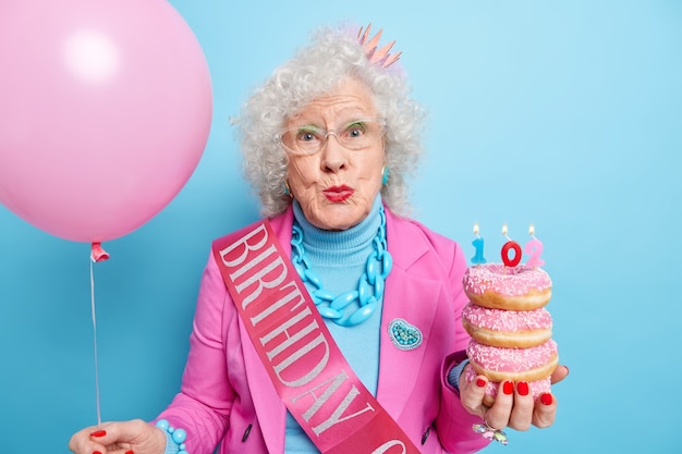 Beautiful curly haired senior woman keeps lips folded enjoys birthday celebration holds pile of delicious doughnuts with candles inflated balloon