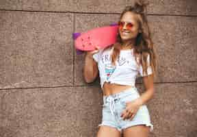 Free photo beautiful cute blond teenager model without makeup in summer hipster white clothes with pink penny skateboard posing near wall on the street