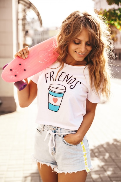 Beautiful cute blond teenager model without makeup in summer hipster white clothes with pink penny skateboard posing on the street background