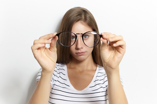 Beautiful cute shortsighted teeage girl in stylish striped t-shirt putting on her first eyeglasses. Vision correction, optics, nearsightedness and myopia concept. Selective focus on spectacles