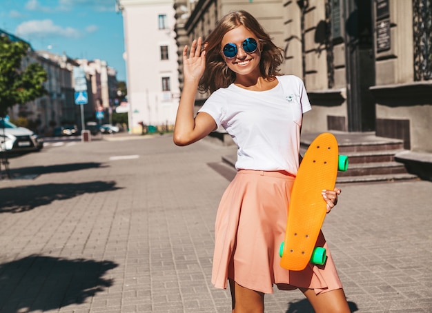 Free Photo beautiful cute smiling blond teenager model in summer hipster clothes with orange penny skateboard posing