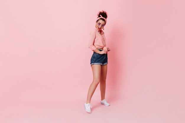 Free photo beautiful dark-haired woman in bandage on her head and lilac glasses posing on isolated space. portrait of girl in white sneakers, shorts and shirt.