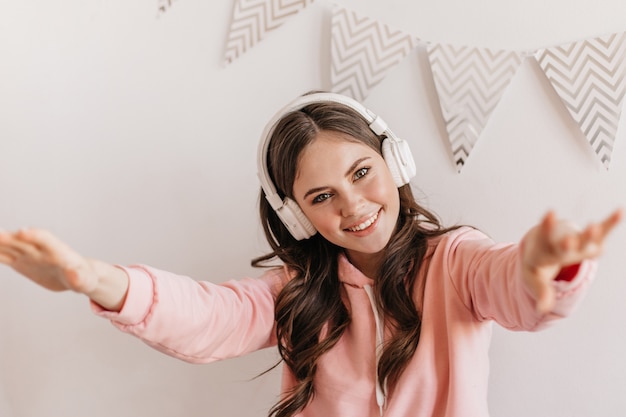 Beautiful dark-haired woman in pink hoodie and massive white headphones posing in bright apartments