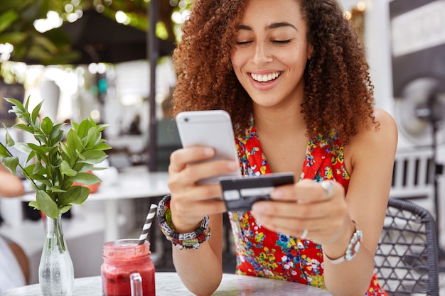 Beautiful dark skinned young female with cheerful expression, holds smart phone and credit card, banks online or makes shopping while sits against cafe interior.