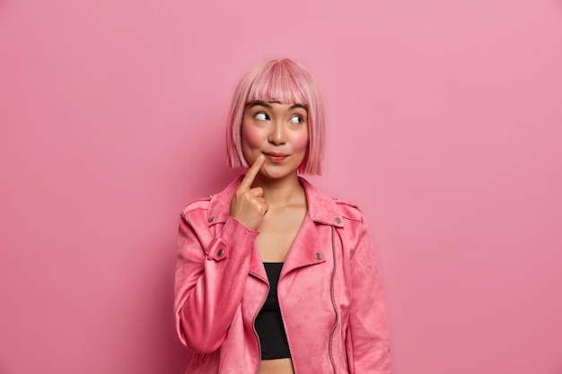 Beautiful dreamy Asian woman looks away with thoughtful expression, keeps finger near mouth, dressed in fashionable outfit has trendy hairstyle pink hair