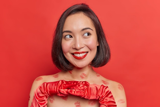 Beautiful dreamy Asian woman smiles gently looks away keeps hands in gloves together decides whether to acceprt invitation and go on date poses against bright red wall has pleasant thoughts