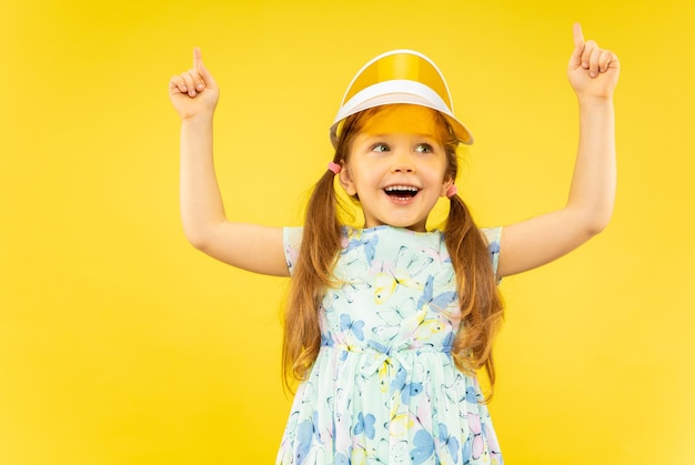 Beautiful emotional little girl isolated. portrait of happy child weard in dress and orange cap pointing up. Concept of summer, human emotions, childhood.