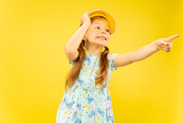 Beautiful emotional little girl isolated on yellow background. Half-lenght portrait of happy child wearing a dress and orange cap pointing up. Concept of summer, human emotions, childhood.