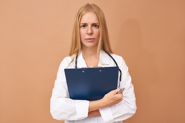 Beautiful experienced young female practitioner in white surgical coat holding clipboard