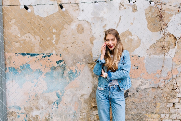 Free photo beautiful fair-haired woman posing sensual in front of the old stone wall. young model talking on the phone outside with grunge wall on the background.