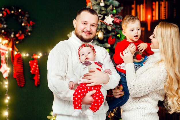 Beautiful family with children in warm sweaters poses before a green wall and rich Christmas tree