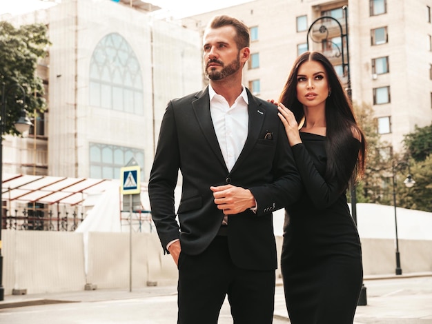 Beautiful fashion woman and her handsome elegant boyfriend in suit Sexy brunette model in black evening dress Fashionable couple posing in the street in Europe Brutal man and his female outdoors