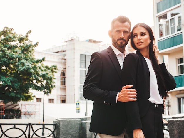 Beautiful fashion woman and her handsome elegant boyfriend in suit Sexy brunette model in black evening dress Fashionable couple posing in the street in Europe Brutal man and his female outdoors