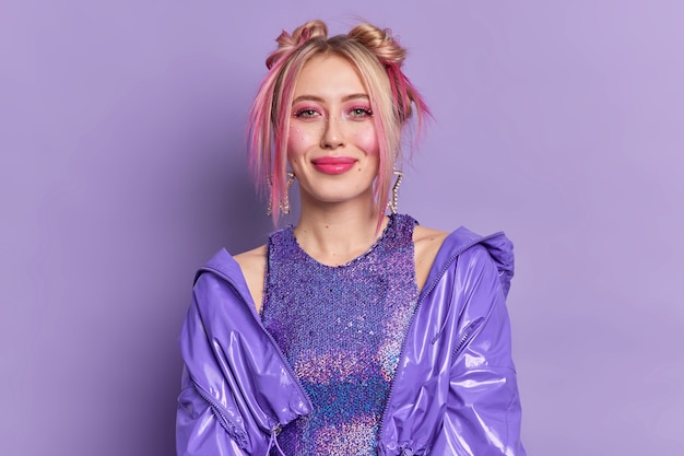 Beautiful fashionable blonde woman with happy expression wears professional makeup has two hair buns dressed in stylish clothes poses against vivid purple wall dresses for disco party.